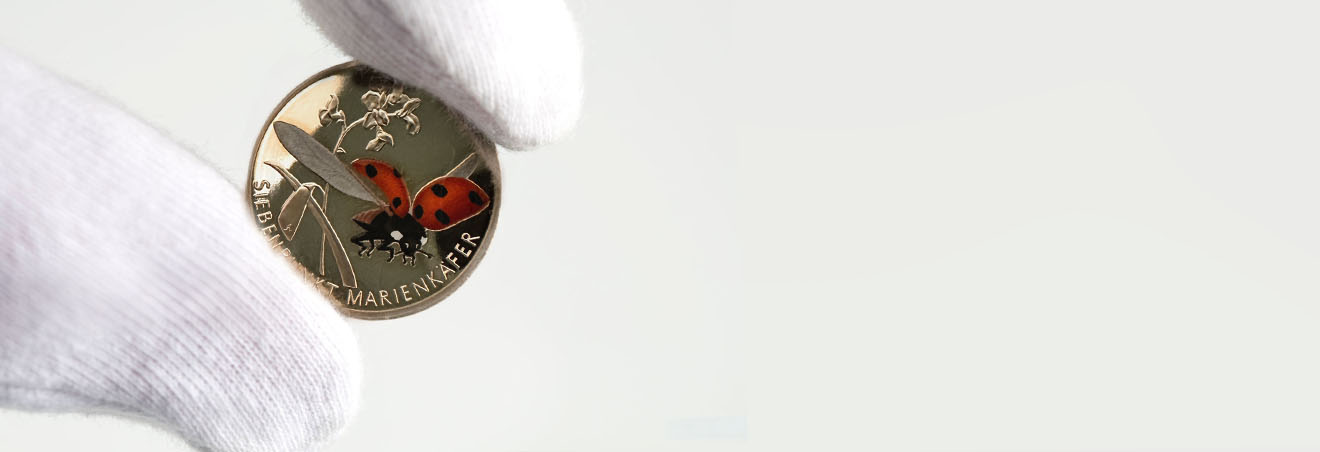 Zwei behandschuhte Finger halten vorsichtig die glänzende 5-Euro-Münze Marienkäfer