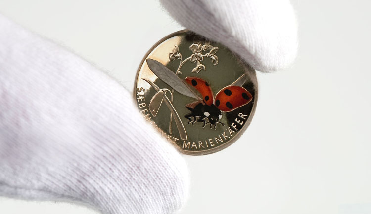 Zwei behandschuhte Finger halten vorsichtig die glänzende 5-Euro-Münze Marienkäfer